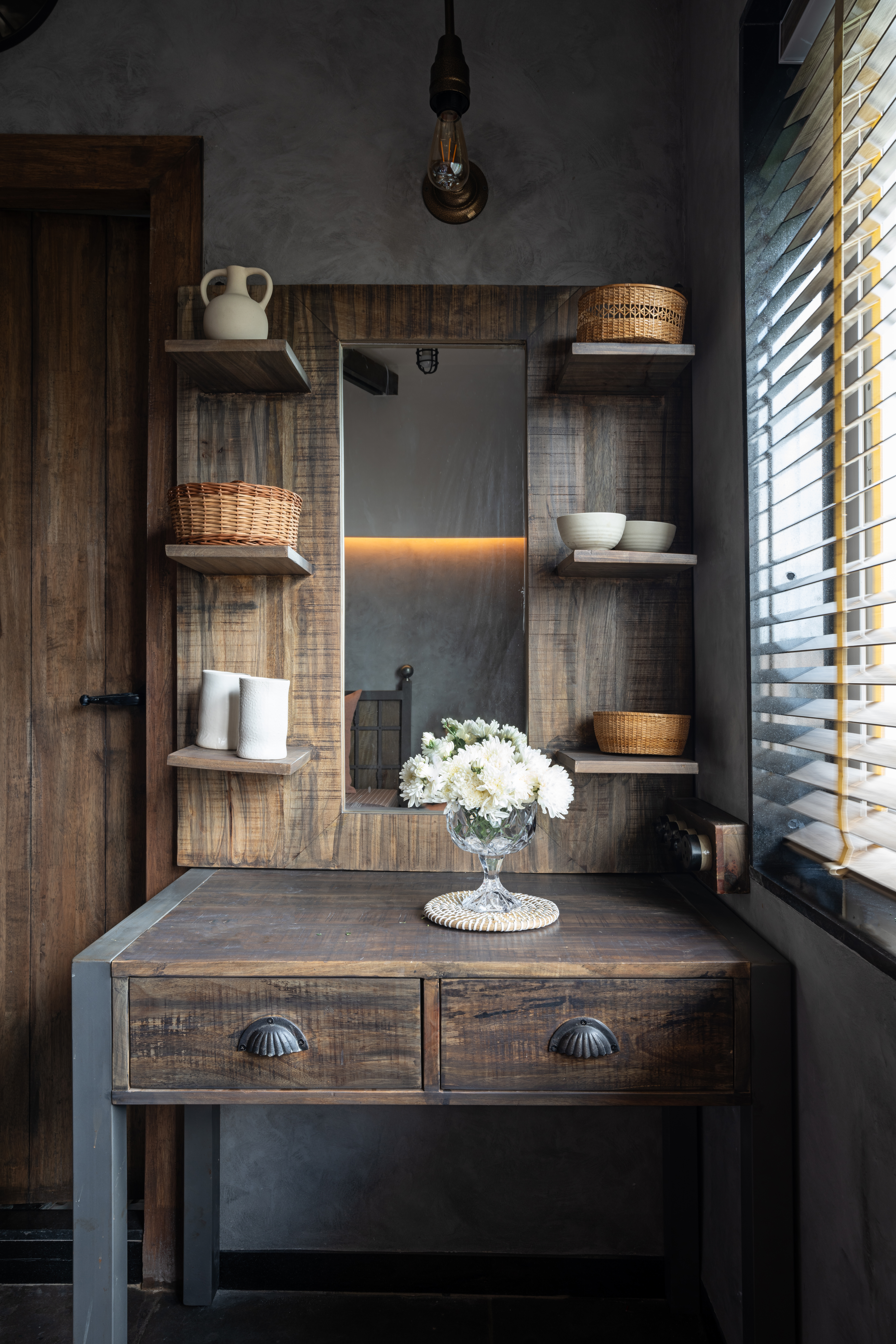 Timber dressing table with cup-handle drawers below a mirror framed by open shelves holding baskets, cream jugs and bowls, with white chrysanthemums in a glass compote on a woven mat, lit by a caged pendant beside a window with timber blinds.